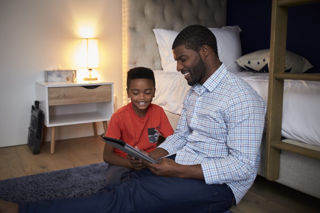 Father With Son Sitting In Bedroom Playing Game On Digital Tablet Together