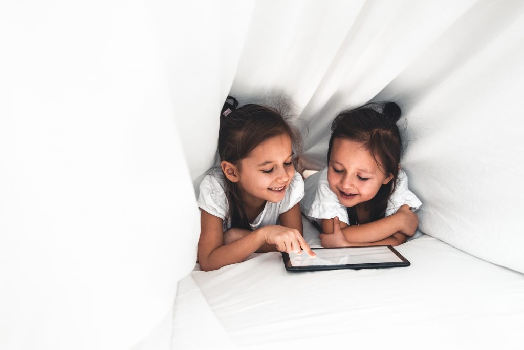 Two beautiful little sisters lying in the bed and look at the screen of a tablet, smart kids using smart technology